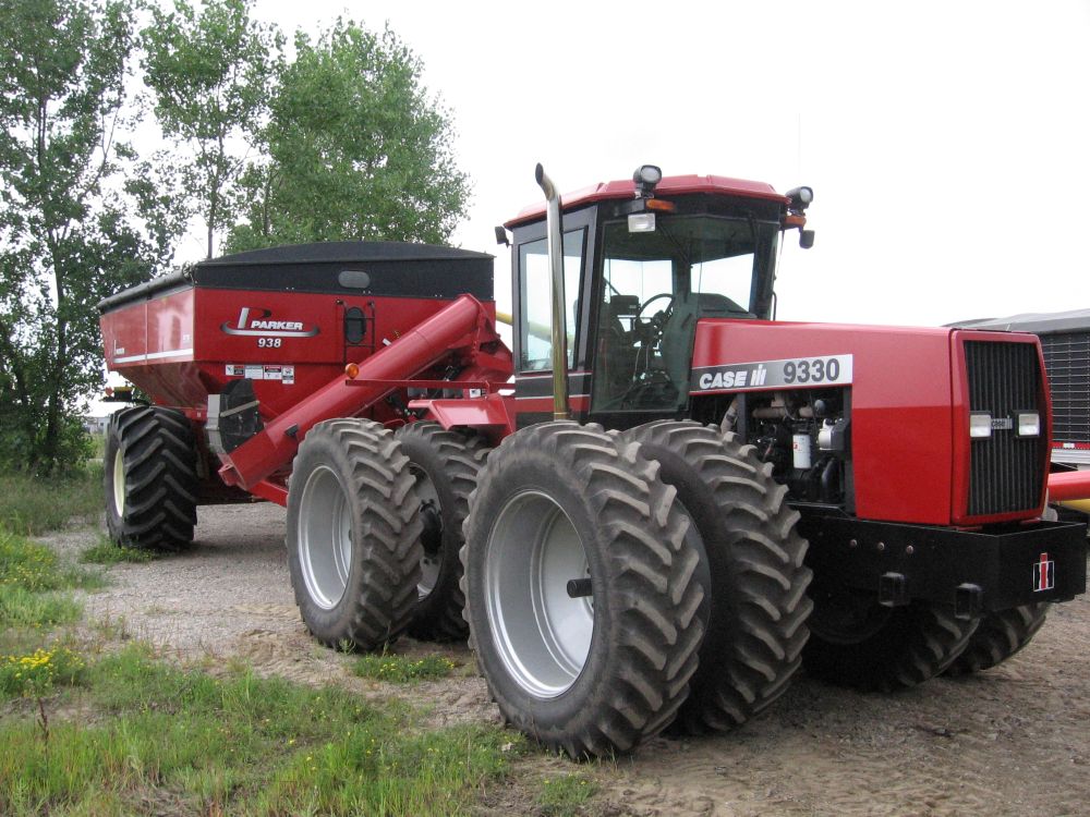 A large red tractor with dual rear and dual front wheels and 1000-bushel grain cart in tow is parked on the sand beside a grassland.