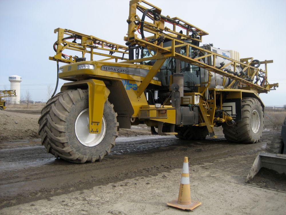 A huge yellow terragator parked on a muddy dirt road in a barren field has three massive tires: two at the rear and one centered in front.