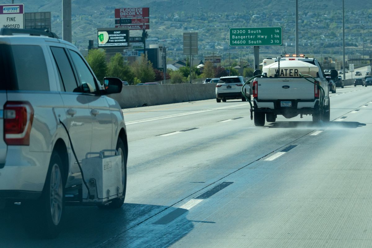 white pickup truck labeled 'WATER' sprays water onto the surface of a Utah highway with tiger-stripe lane markings. Behind that truck, another white utility vehicle equipped with a retroreflectometer gauges the reflectivity of the wet lane markings.