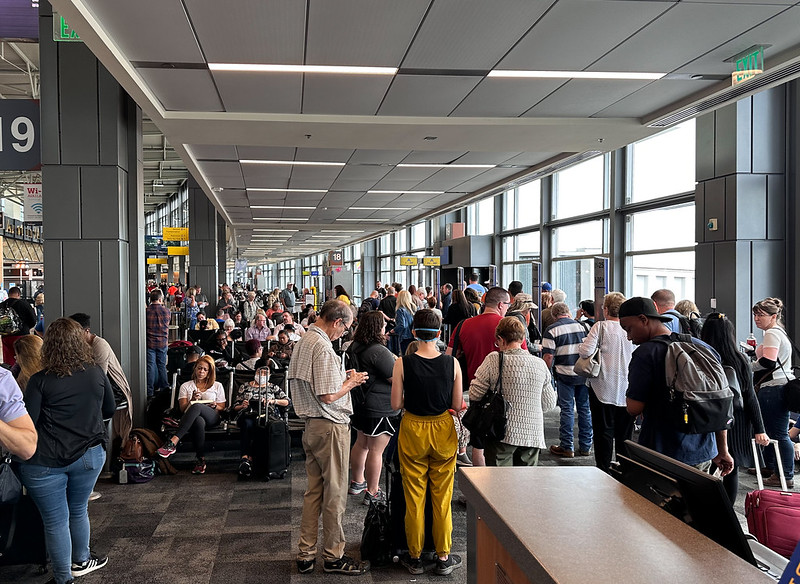 Passengers at the gate of Austin-Bergstrom International Airport in Texas are standing, sitting, or checking their phones, with many carrying luggage or backpacks.