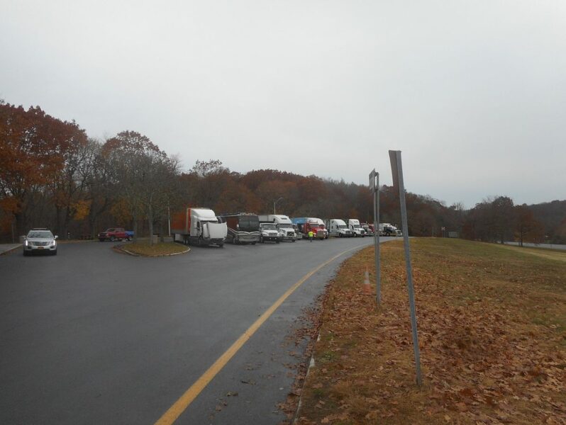 Several trucks and a few cars are parked at a rest area in Bedford, New York, with autumn foliage and bare trees in the background.