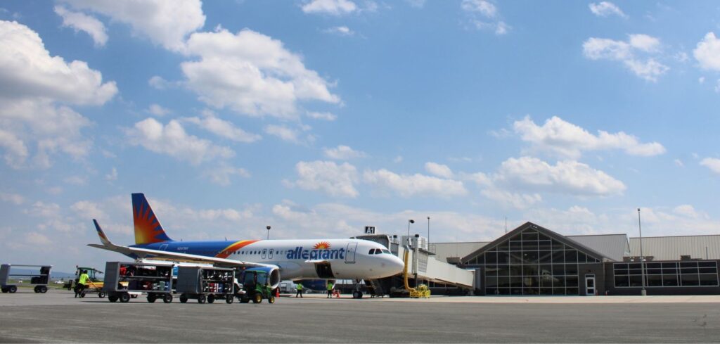 A small commercial airplane parked at a gate connected to the terminal at Hagerstown Regional Airport in Maryland via a jet bridge. Two open-sided runway baggage carts full of suitcases sit next to the plane while several ground crew members in yellow safety vests work nearby on the mostly empty tarmac.