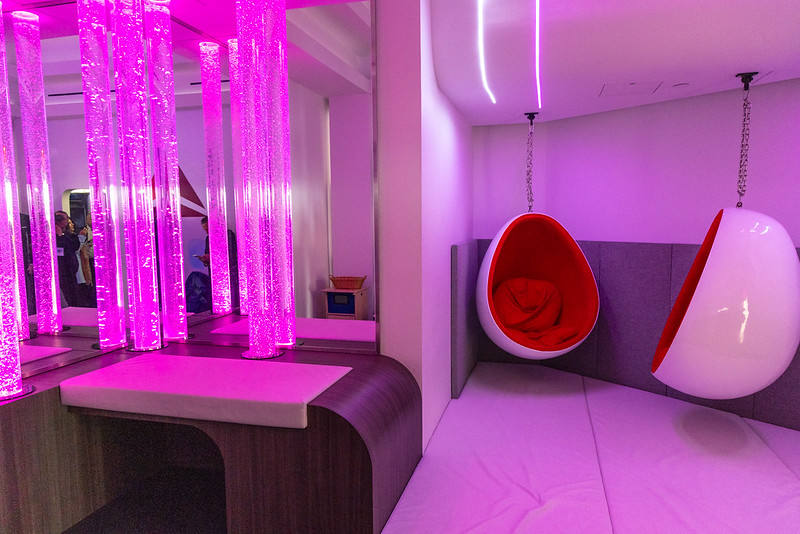 A vibrant multisensory room at New York's LaGuardia Airport features two modern egg-shaped hanging chairs with red interiors suspended in a corner with soft padded flooring. The other side of the room shows a set of illuminated bubble tubes emitting pink light.