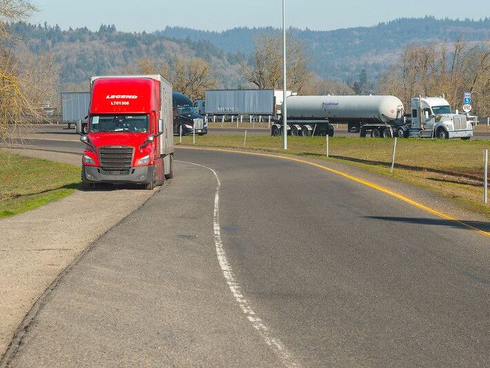 A red semi-truck is parked on the offramp shoulder on Interstate 5 in Jefferson, Oregon. In the background are a white tanker and a freight truck parked on the highway shoulder.