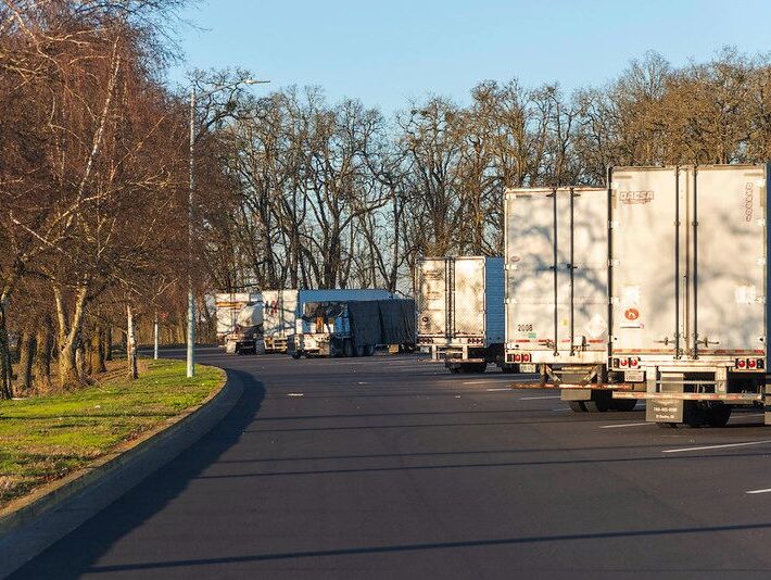 Multiple semi-trucks and trailers parked at an Oregon rest area with wide, paved road with trees on either side.