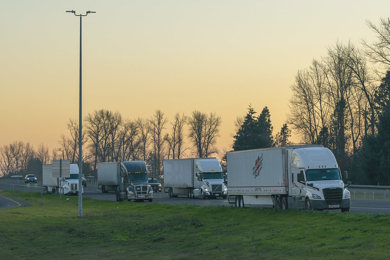 An Oregon highway scene shows a line of semi-trucks and trailers parking on the shoulder at sunset.
