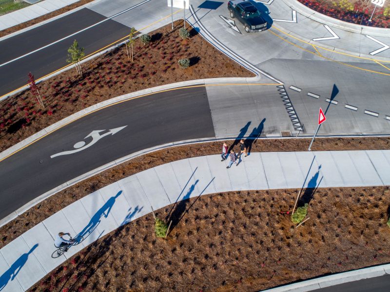 Aerial view of a roundabout with a shared-use path running alongside it shows a bicyclist riding on the path while pedestrians walk nearby. A vehicle navigates the roundabout, and yield signs are posted at pedestrian crossings.