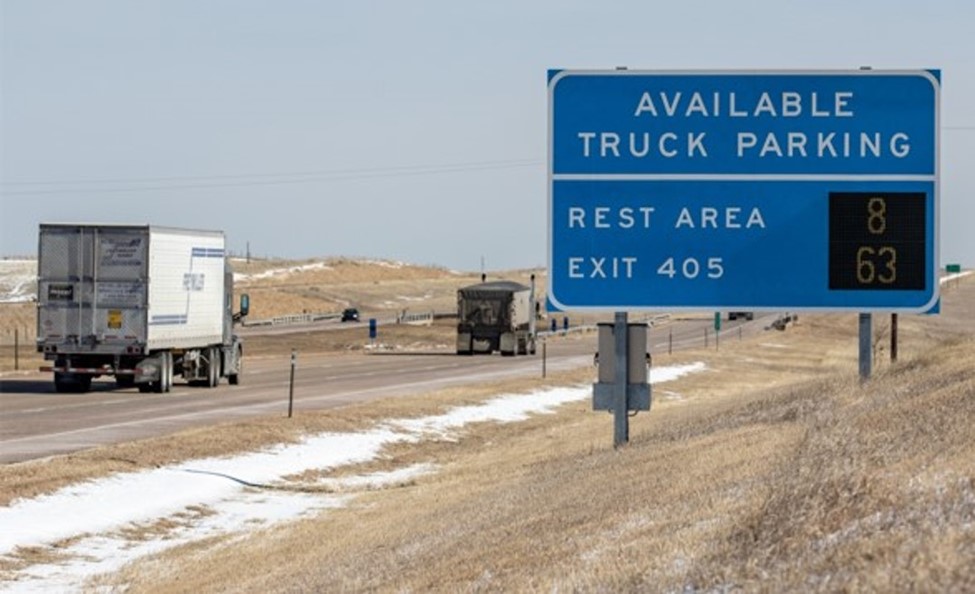 Highway view of freight trucks passing a large blue sign with electronic numbers that reads 'Available Truck Parking' and 'Rest Area: 8; Exit 405: 63.'