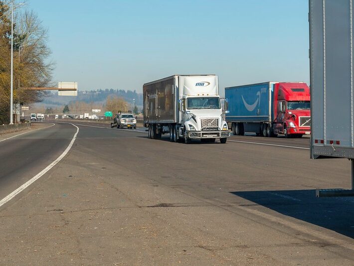 Highway scene shows a white freight truchk parked on the wide junction between an offramp and the busy main road in Oregon. The scene is set on a clear day with trees lining the left side and hills visible in the background.