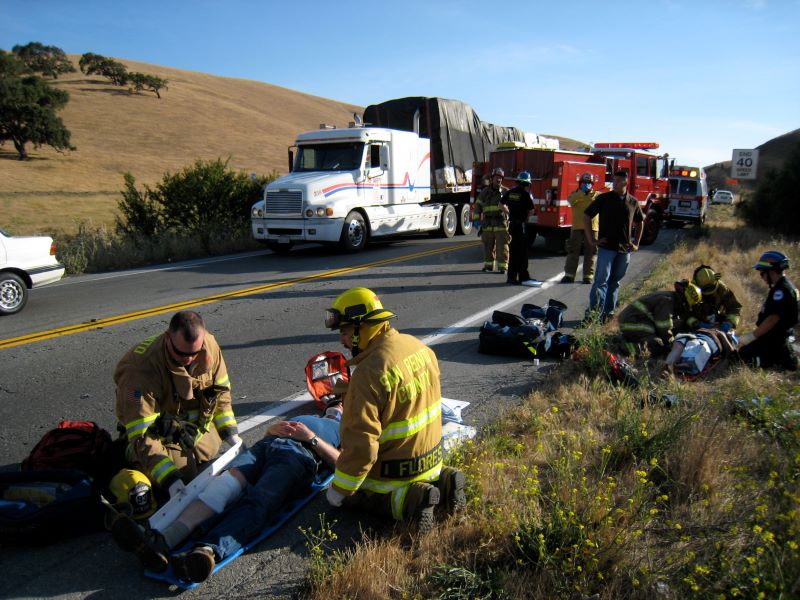 Firefighters and medics treat injured people on the roadside after a crash involving a truck, with fire engines and crews nearby.