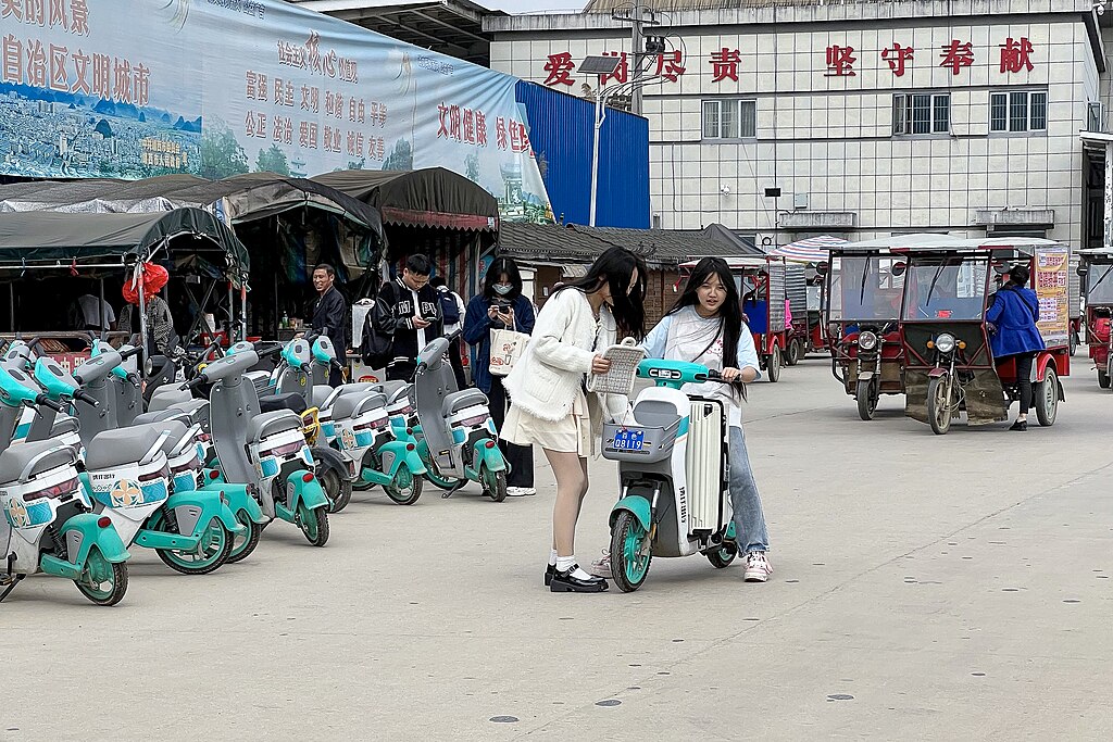 Two young women use a shared e-bike in Jingxi, China, while rows of rental e-bikes are parked nearby and people move through the busy market area.