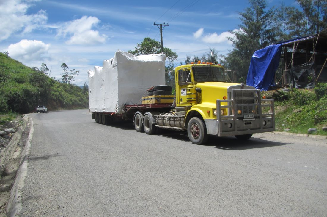 A yellow truck with a flatbed trailer is on a paved road carrying large wrapped mining equipment, with additional smaller equipment loaded on the trailer.