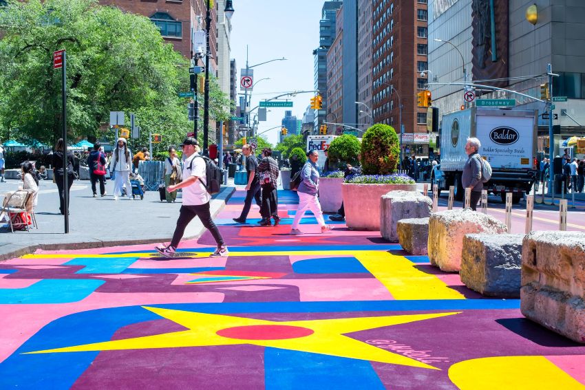 A New York City street has colorful painted pavement with stone barriers and planters used for hardened daylighting as pedestrians cross the area.