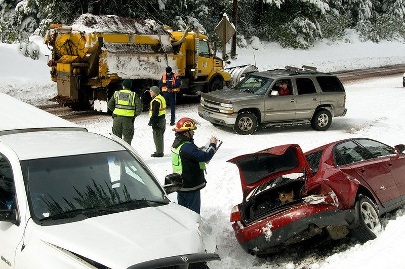 Police and emergency workers respond to a snowy road crash in Oregon involving multiple vehicles, while a snowplow truck clears the road in the background.