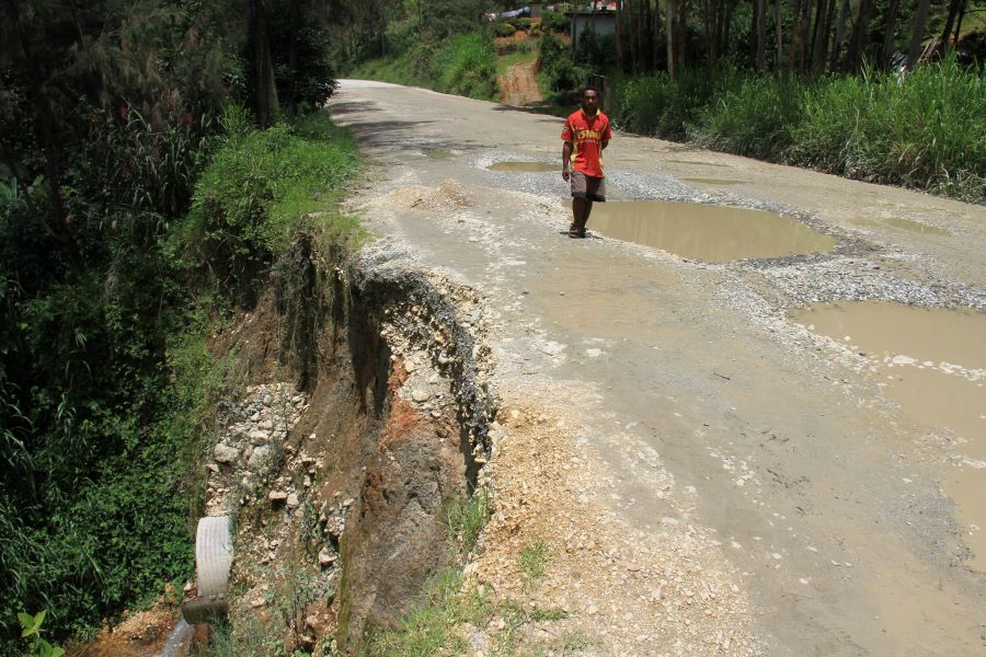 A damaged unpaved rural road with large potholes and standing water is partly eroded at the edge while a person walks carefully along the surface.