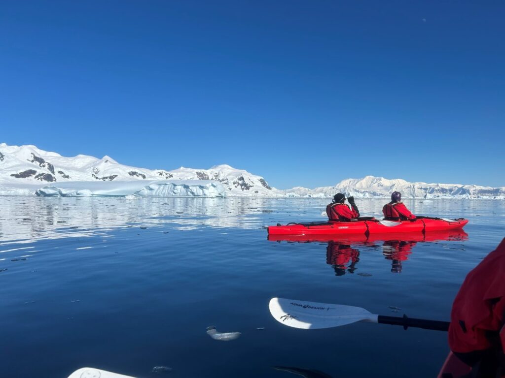 Two people in red parkas and black hats sit in a red, two-person kayak in calm water off Antarctica with snowy peaks and icebergs in the background. The paddle and left elbow of a red-jacketed kayaker is in the far right corner of the foreground.