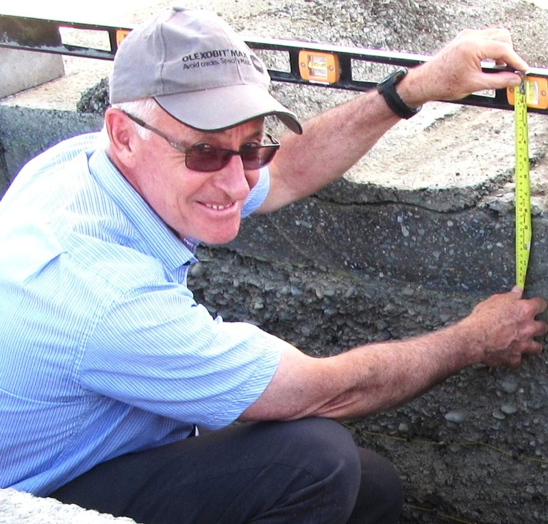 David Jones, in a blue short-sleeved shirt and cap, looks up from the roadside trench where he is using a yellow measuring tape and a level to measure pavement layers.