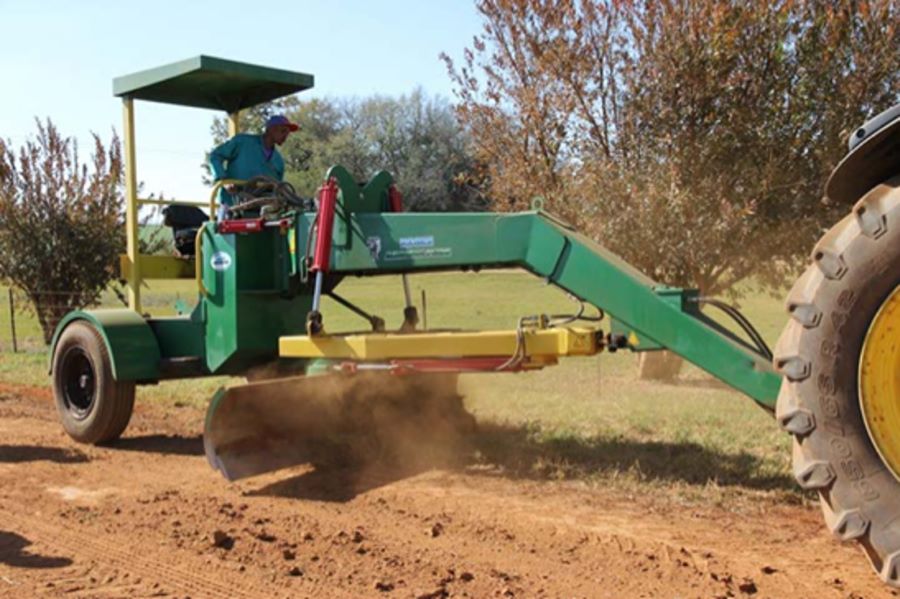 A green grader with a front blade is pulled by a tractor while a seated operator levels the surface of an unpaved rural road.