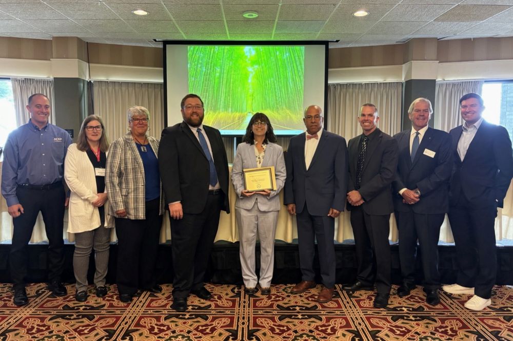 A group of nine people stands indoors as A C R P Manager Marci A. Greenberger at the center holds a 2025 Award of Excellence presented by the Michigan Aeronautics Commission and Michigan Department of Transportation.