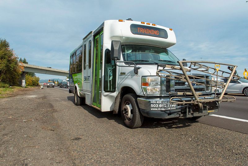 A bus with a front-mounted bike rack and visible training signage above the windshield drives in the breakdown lane of a busy highway.