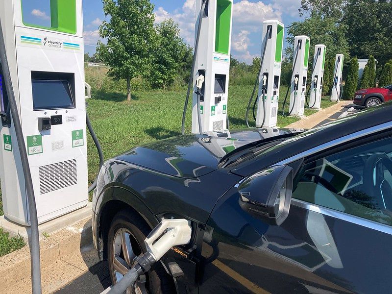 A car charges at a row of electric vehicle fast charging stations in a parking area beside grass and trees.