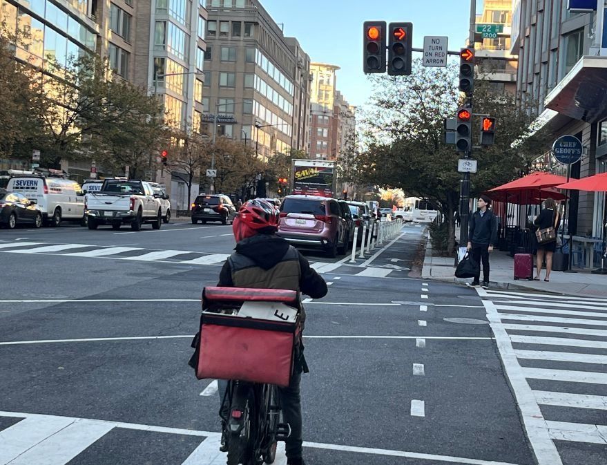 A cyclist on an electric bicycle with a large delivery bag waits in the bike lane at a busy intersection while the traffic light shows red for vehicles, bikes, and pedestrians.