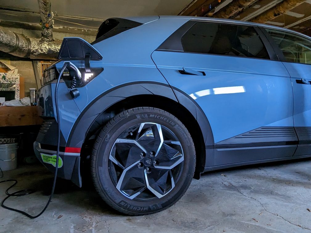 An electric vehicle sits in a garage with a charging cable plugged into its port near the rear wheel.