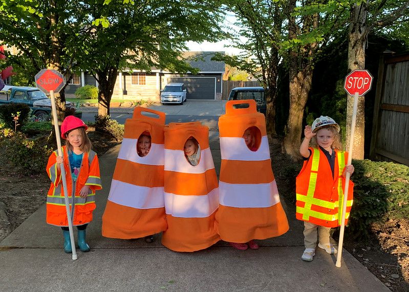 Children dressed as traffic cones and safety workers stand on a sidewalk holding stop signs and wearing bright safety gear.