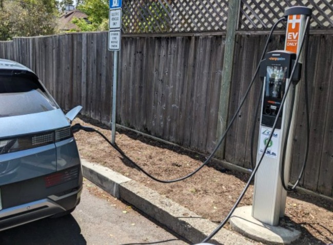 A parked electric vehicle connects to a Level 2 charging station with a long cable beside a wooden fence near a library parking area.