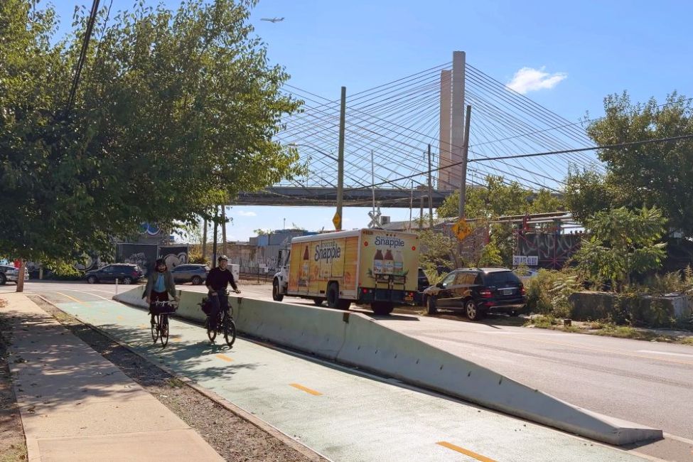A raised median barrier separates a bike lane from the road as cyclists ride beside traffic near a large cable stayed bridge.