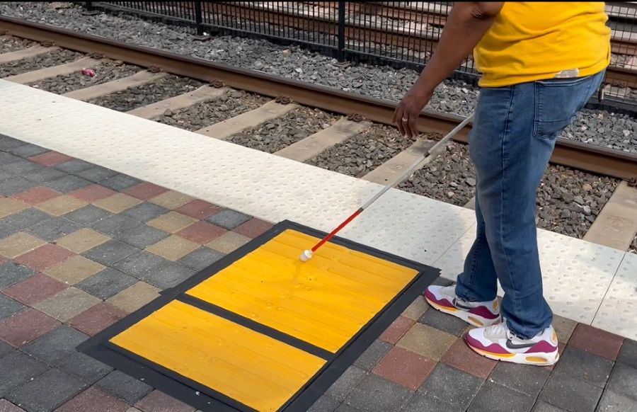 A person uses a white cane to locate a yellow tactile path beside a train platform edge.