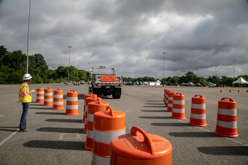 A work-truck driver competing on a large obstacle course maneuvers backward between a narrow lane of large orange barrels while a judge wearing a hard hat and safety vest scores the skills.