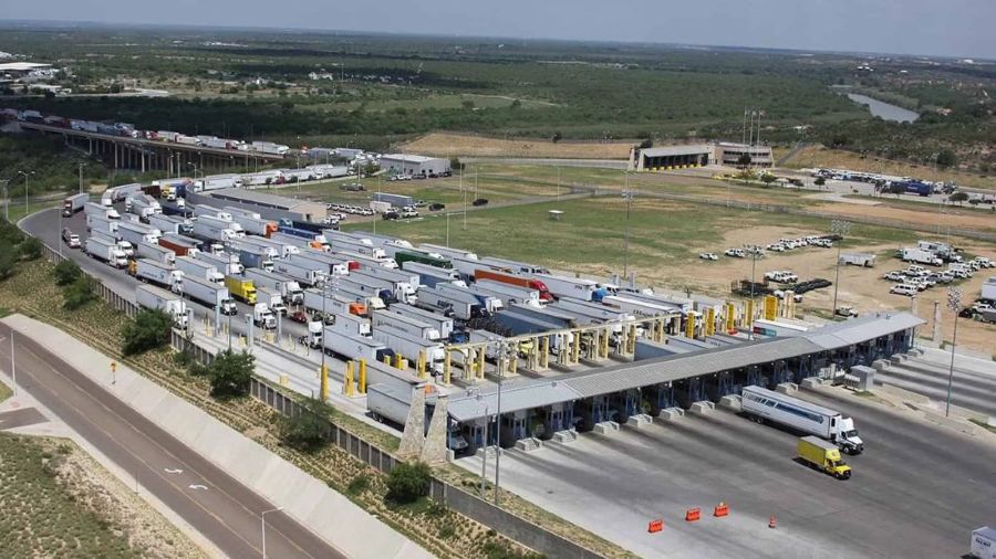The aerial view of the World Trade International Bridge shows many trucks lined up in multiple lanes. The trucks wait at a series of inspection booths with canopies and equipment. The lanes extend toward the bridge structure in the distance. A few trucks move through open lanes while many others remain stopped. The area around the bridge includes paved roads, open land, parking areas, and scattered buildings. Trees and a river appear in the background along with wide stretches of land.
