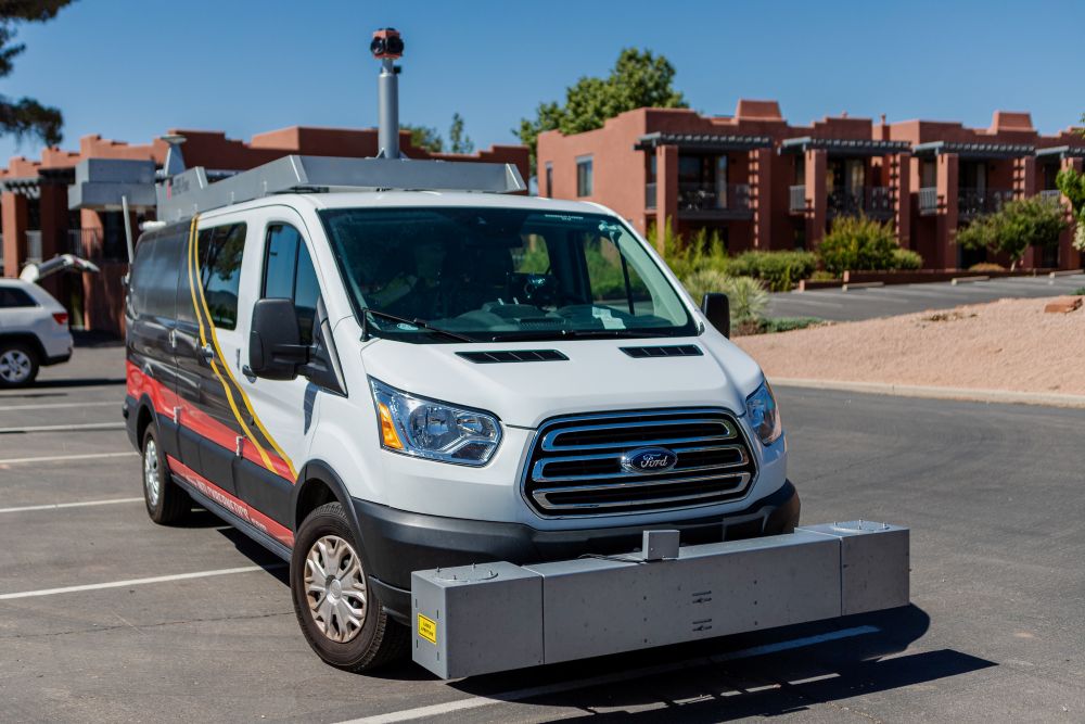 An AI-enabled road survey vehicle with a rectangular box-shaped device mounted on the front bumper moves on a road near buildings.