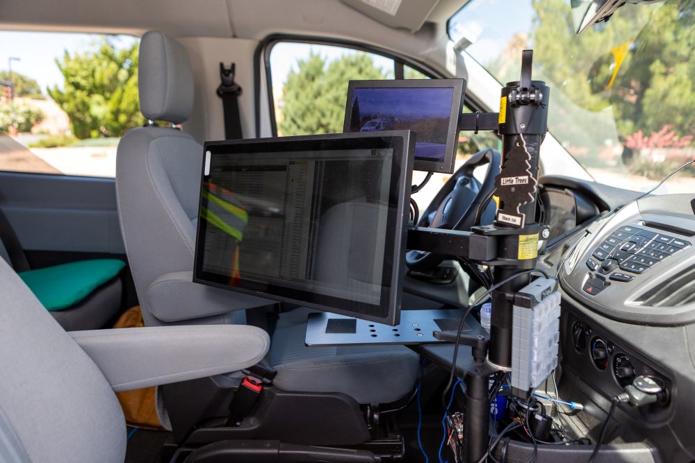 The inside of an AI-enable road survey vehicle shows screens and devices mounted on a frame near the passenger seat. A keypad is on the dashboard.
