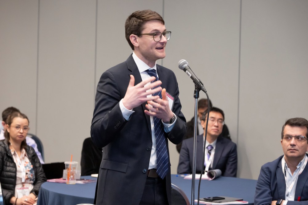 Alex Marach, in formal wear, stands near a microphone and speaks with people seated behind him.