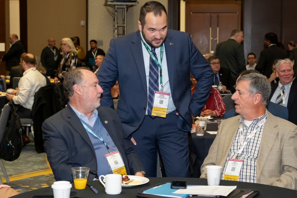 Kevin Keller, Seçkin Özkul, and Keith Bucklew, in formal wear with I D cards, gather around a table in an indoor hall and talk, with beverages on the table.