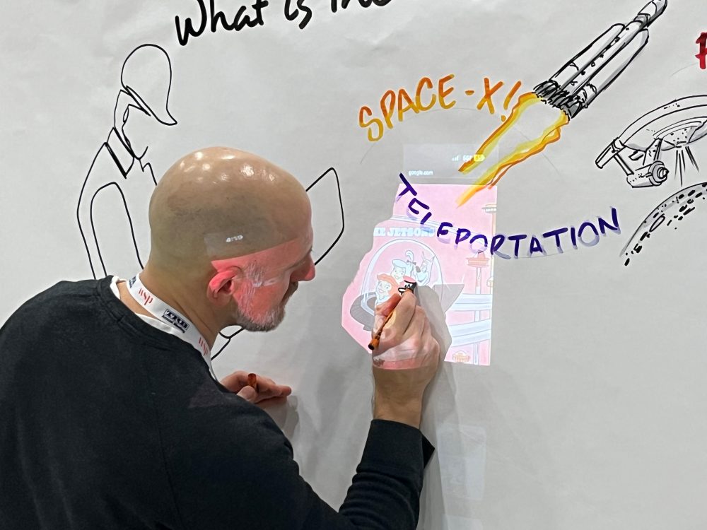 Brian Tarallo traces the Jetsons in their flying car on the wisdom wall, adding sketches and text including Space X, teleportation, a rocket, and other illustrated elements.