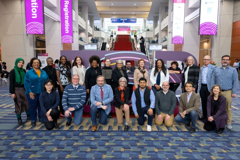 Cooperative Research Programs staff pose with large T R B letters behind them in the lobby of the Washington Convention Center during the T R B Annual Meeting. Some stand in the back row and some kneel in the front row. Stairs and banners are in the background.
