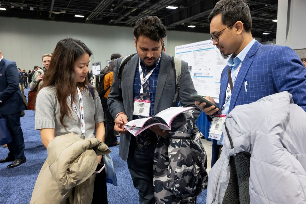 Three students wearing T R B Annual Meeting I D cards look at the program while standing in the poster session hall holding jackets. phones, and a backpack.