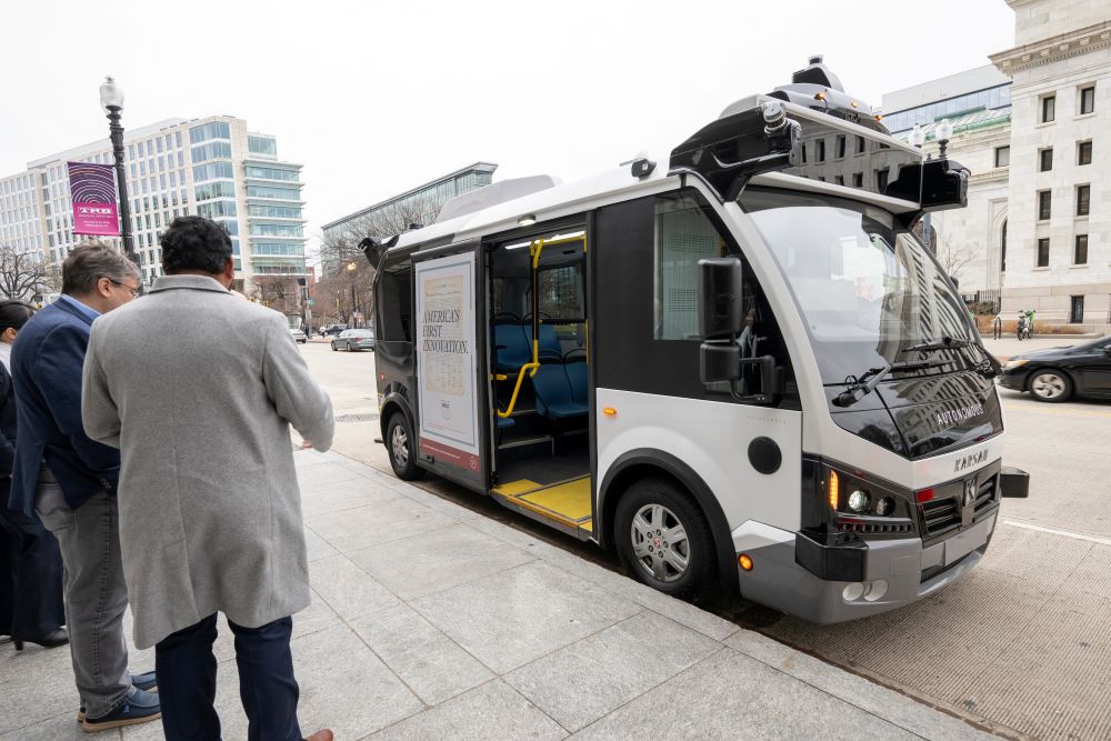 A small autonomous bus is parked at a curb outside the Washington Convention Cetner with its door open and floor level with the curb. Two people stand near the bus and look toward it.