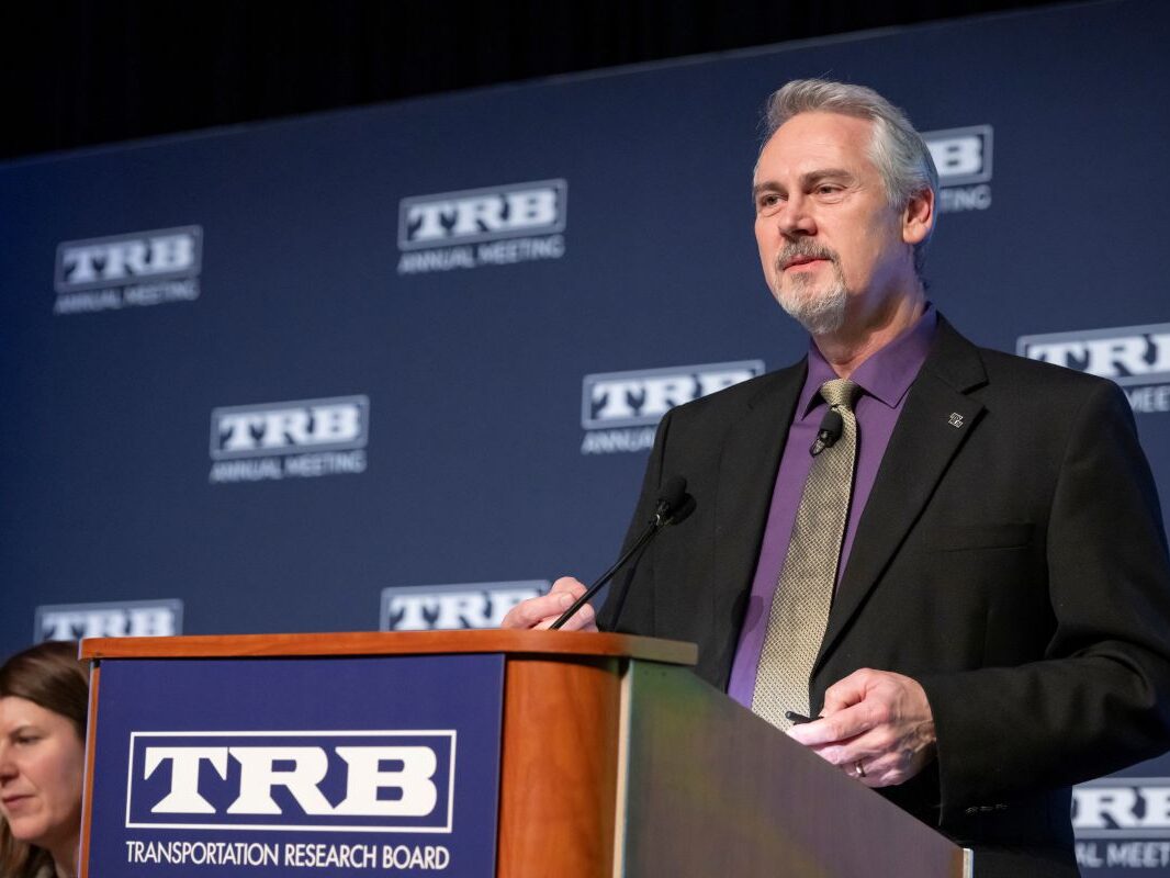 David Harkey Deen Lecture David Harkey stands at a lectern with T R B Transportation Research Board text on it and delivers the Deen Lecture as T R B Executive Director Victoria Sheehan sits and listens on the side.