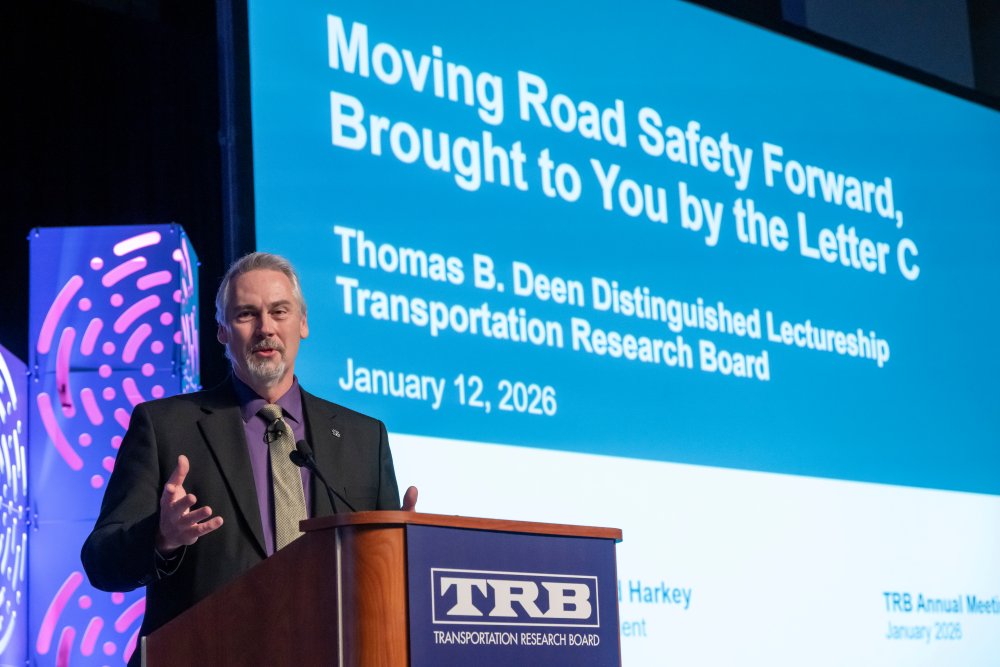 David Harkey speaks at a lectern while a presentation screen behind him reads Moving Road Safety Forward, Brought to You by the Letter C, which is the title of his Thomas B. Deen Distinguished Lectureship at the 2026 T R B Annual Meeting.