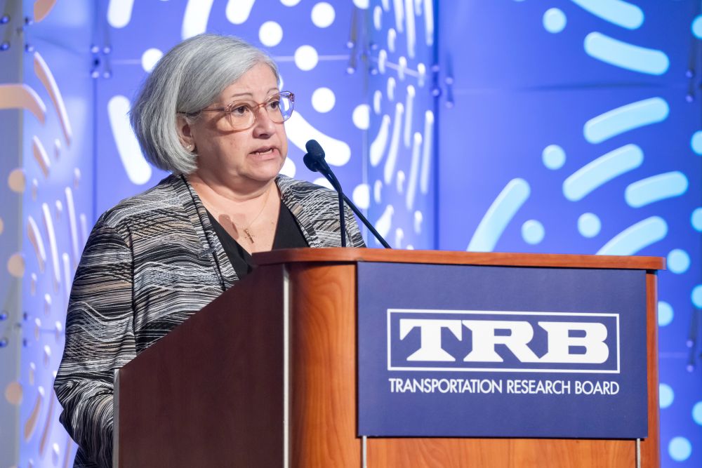 Diane Gutierrez-Scaccetti stands at a podium and speaks into a microphone, with text on the podium reading T R B Transportation Research Board.