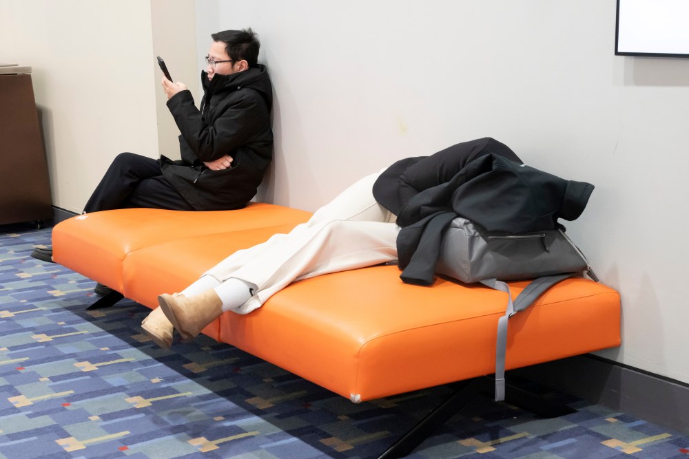 Two attendees rest on a bench. One person sits and uses a phone. The other person lies down with coat-covered head resting on a backpack.