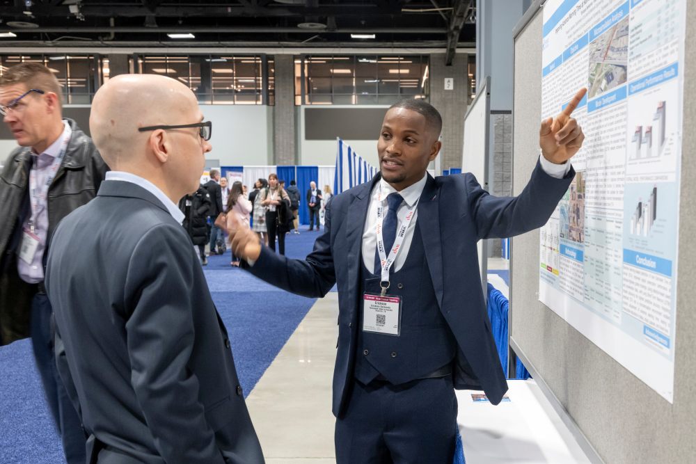 Erickson Senkondo, in formal wear, points at his poster and explains to Joffrey Lauthier, in formal wear, in an indoor hall.