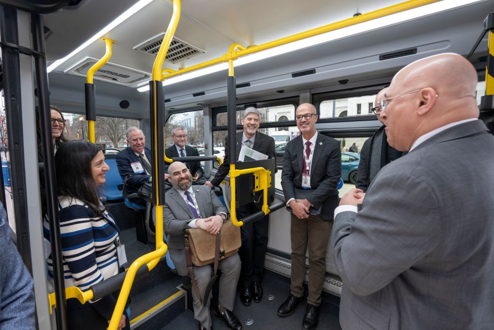 T R B Executive Committee members and other V I P attendees in formal attire stand and sit inside an autonomous bus and talk. Handrails and seats are inside the bus.