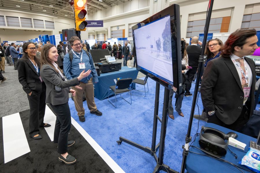 Phea Padgett gestures toward a monitor displaying a street scene while Kristin Kersavage and Bryan Katz stand nearby among exhibit booths and equipment.
