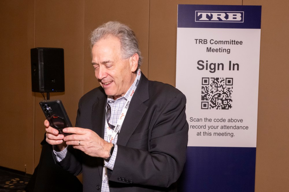 Fred Wagner holds a phone and smiles. A sign behind reads T R B Committee Meeting, Sign In with a Q R code. Scan the code above to record your attendance at this meeting.