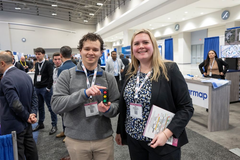 Lance Alex Kloefkorn holds a rubber traffic light while standing with Katryna Pruitt, who holds a file, as they pose among exhibit booths and attendees.